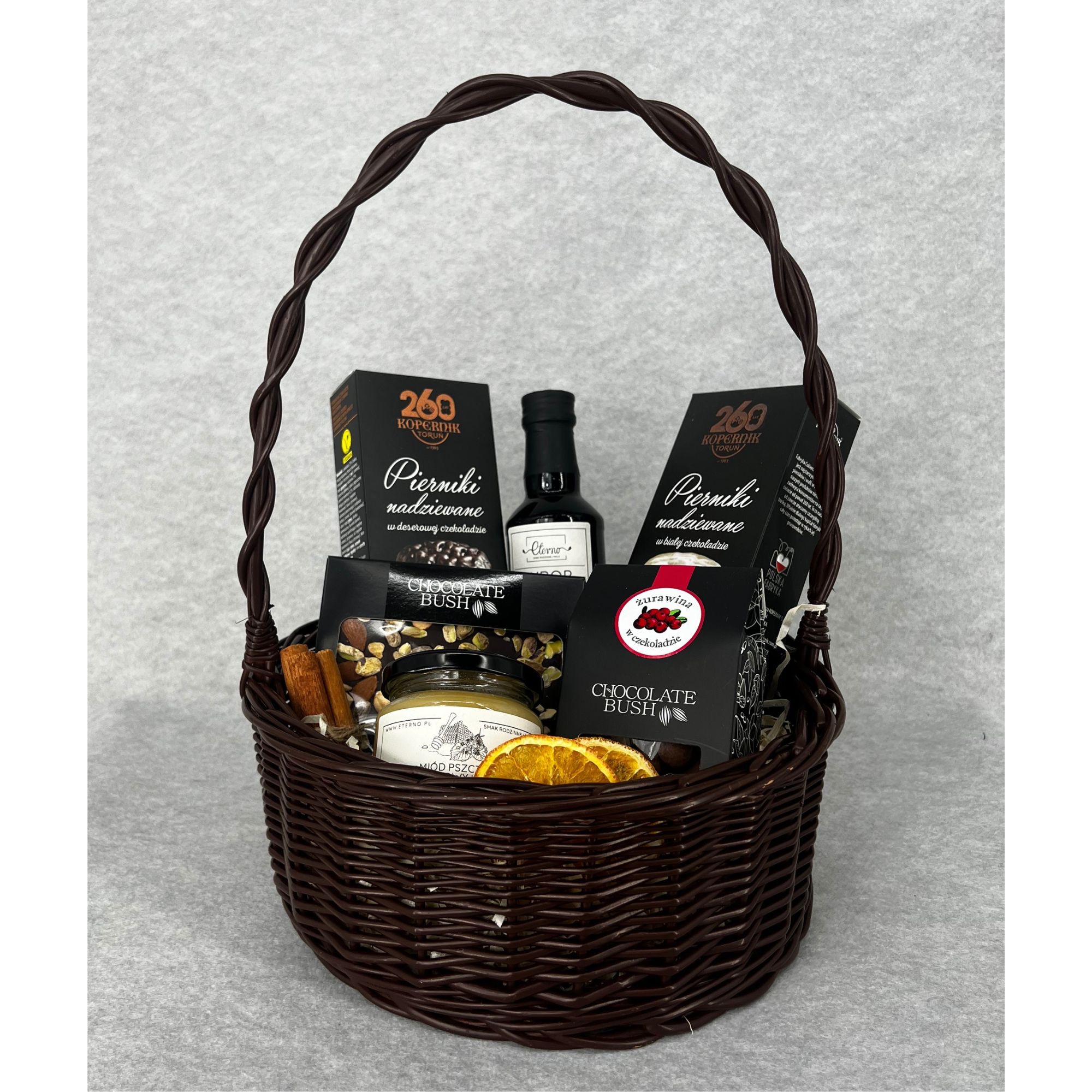 Ciemnobrązowy kosz wiklinowy wypełniony różnorodnymi produktami spożywczymi, w tym piernikami marki Kopernik, miodem pszczelim w słoiku, butelką z etykietą 'Eterno' oraz czekoladami Chocolate Bush,...