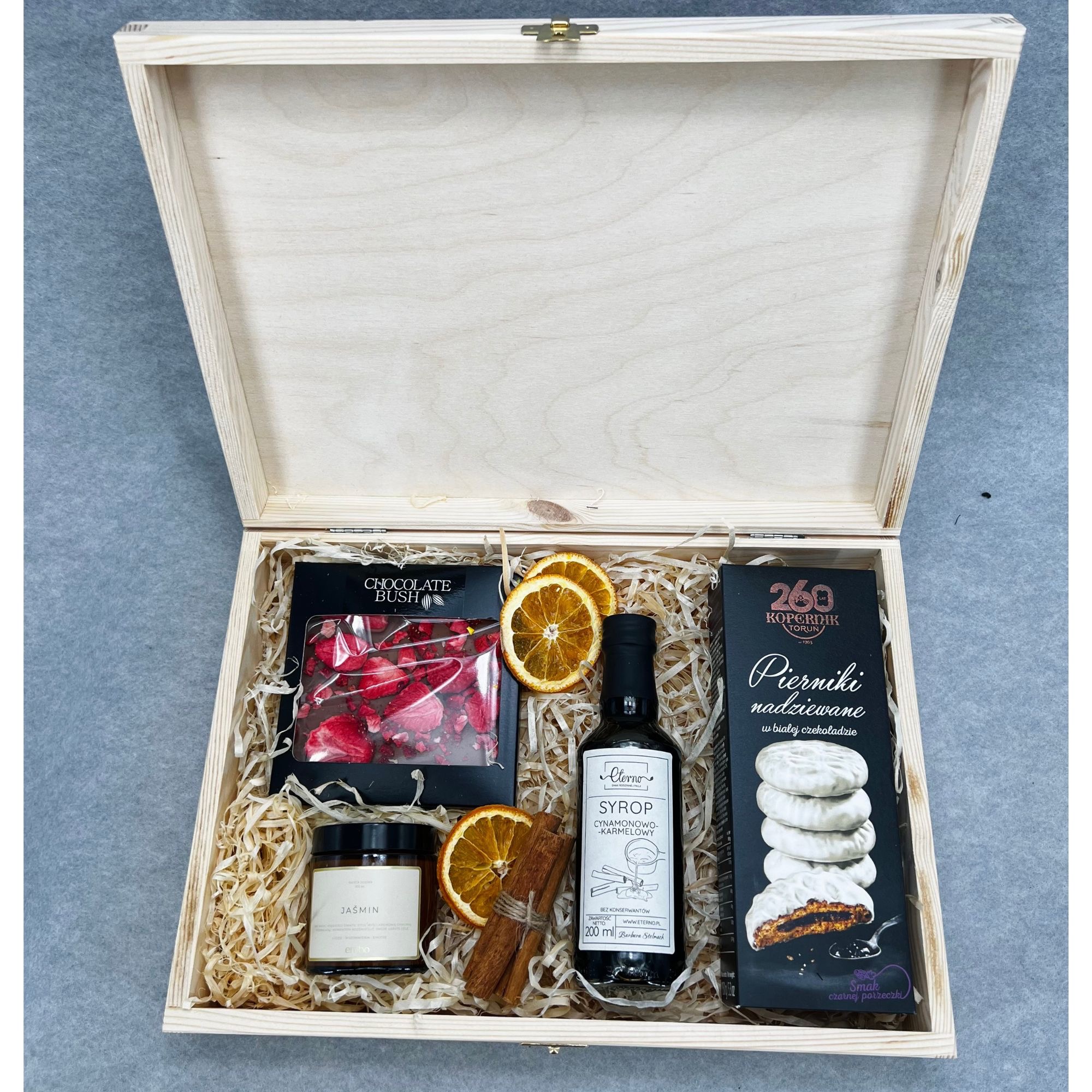 Otwarty drewniany kufer wypełniony prezentami: czekolada z truskawkami, syrop cynamonowo-karmelowy, pierniki w białej czekoladzie, świeca zapachowa Jaśmin, plasterki pomarańczy i laski cynamonu...