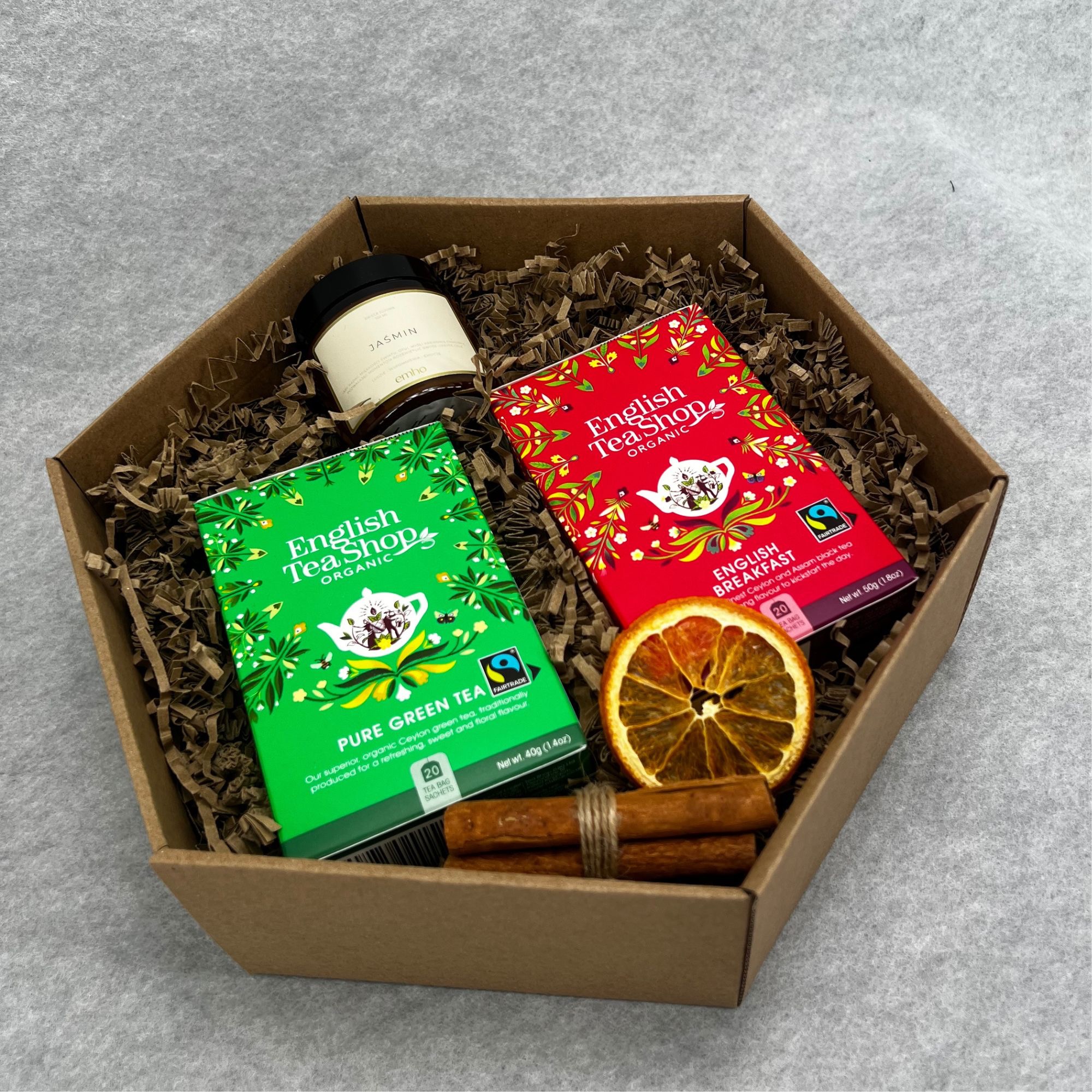 Otwarty kartonowy sześciokątny pojemnik wypełniony brązowym wypełniaczem, zawierający herbatę English Tea Shop (zieloną i czerwoną), słoik z etykietą Jasmin oraz suszoną pomarańczę i cynamon.