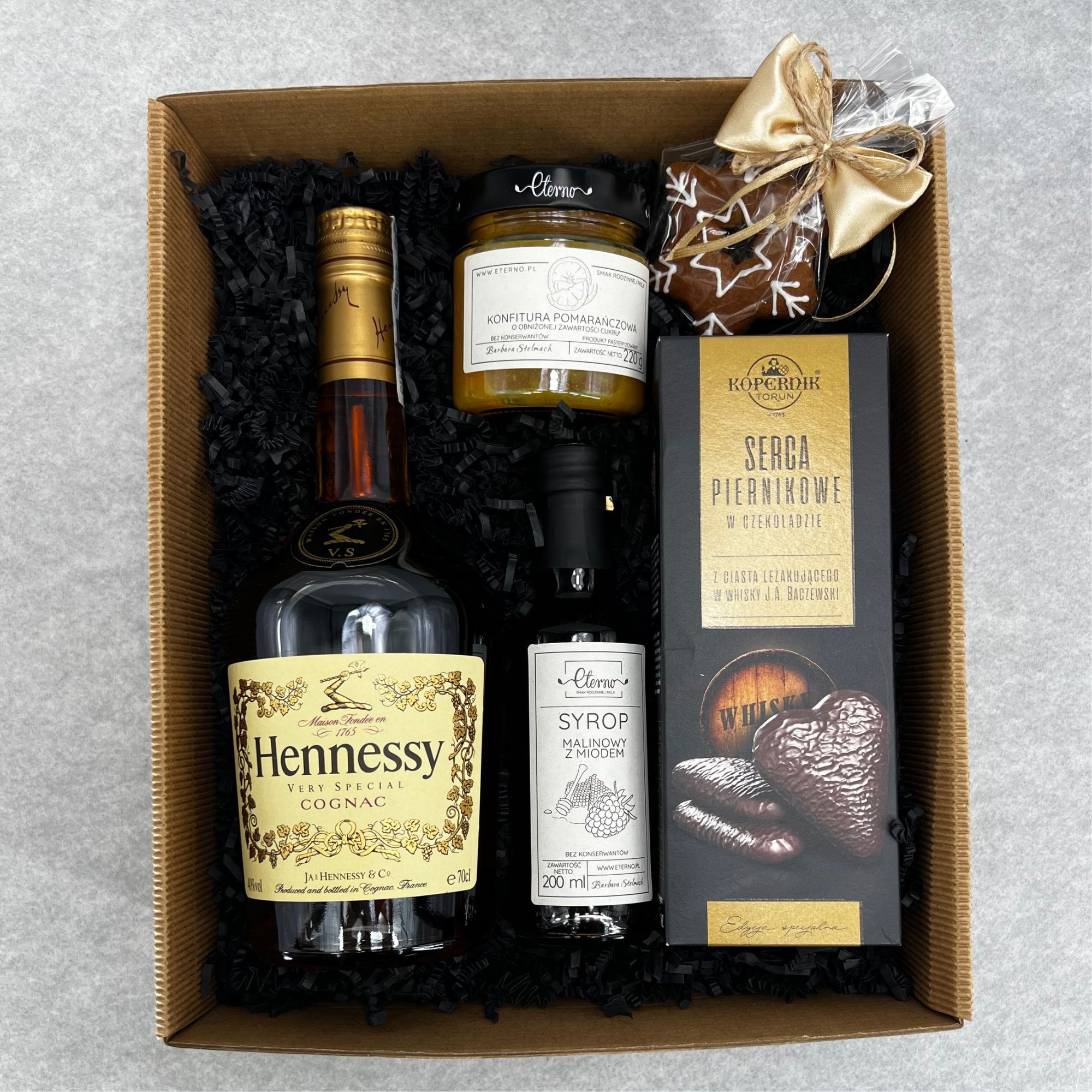 Zestaw upominkowy w kartonowym pudełku wypełnionym czarnym wypełniaczem, zawierający koniak Hennessy, konfiturę pomarańczową, syrop malinowy z miodem, pierniki w czekoladzie w kształcie serc...