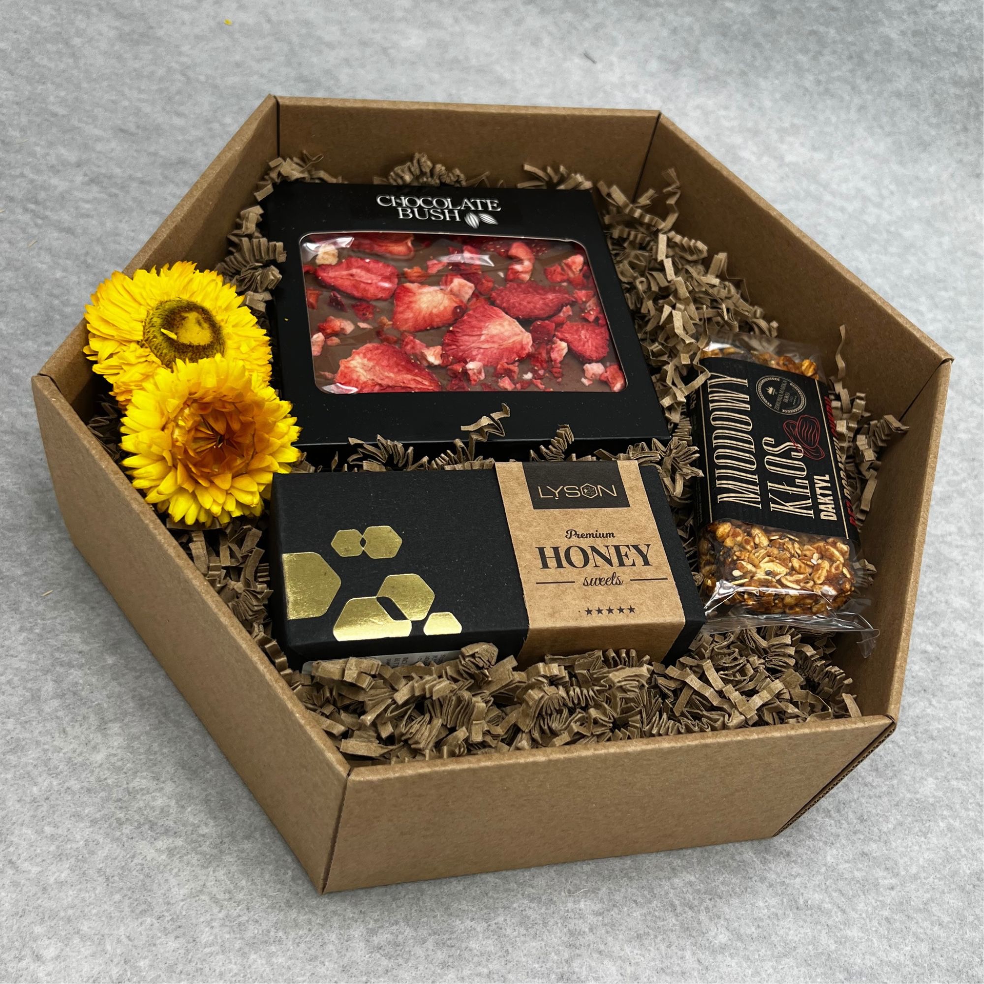 Hexagonalny kartonowy box wypełniony brązowym wypełniaczem, zawierający czekoladę z truskawkami w czarnym opakowaniu, miód w czarnym pudełku z etykietą i batonik daktylowy w przezroczystym...