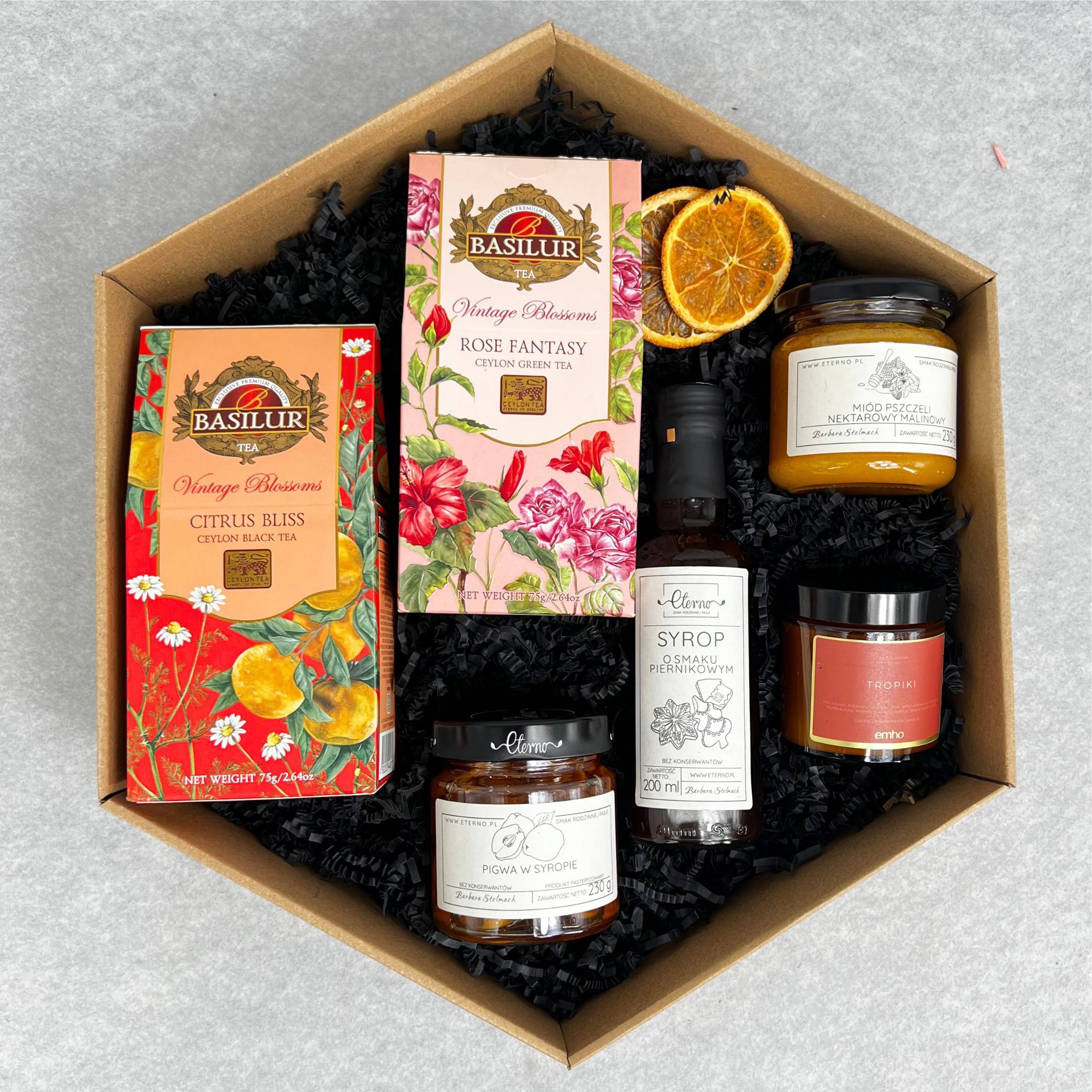 Otwarty, sześciokątny kartonowy box z czarnym wypełniaczem, zawierający dwa opakowania herbat Basilur Vintage Blossoms (Citrus Bliss i Rose Fantasy), słoiczek miodu malinowego, słoiczek pigwy...