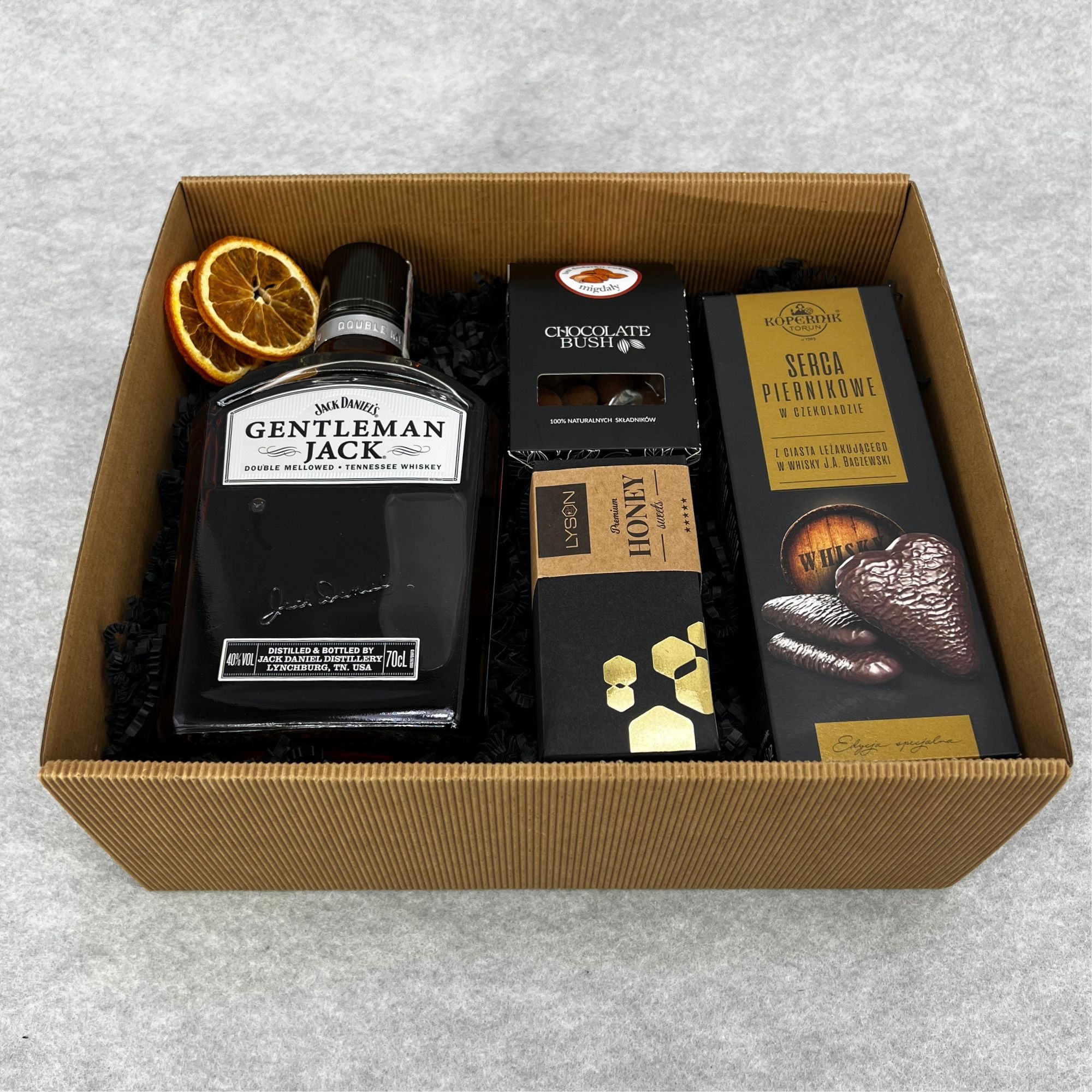 Elegancki upominek w tekturowym pudełku: butelka whisky Gentleman Jack, czekoladki Chocolate Bush, miód Lyson i piernikowe serca Kopernik, udekorowany suszonymi plasterkami pomarańczy.