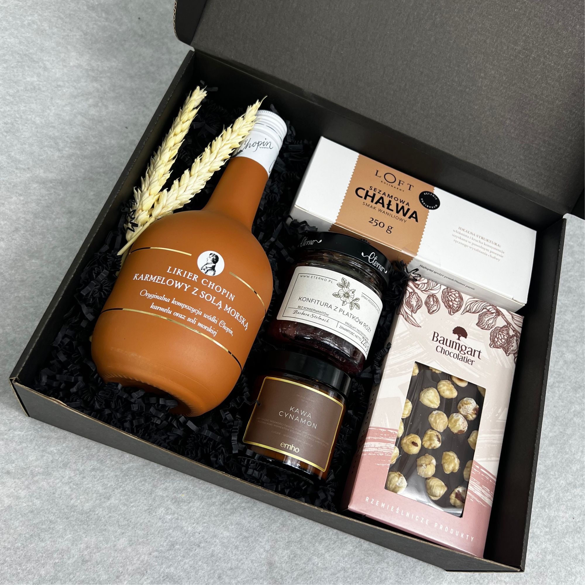 Czarny kartonowy box wypełniony czarnym siankiem, zawierający butelkę likieru karmelowego Chopin, słoik konfitury z płatków róży, puszkę kawy cynamonowej, chałwę sezamową i czekoladę z orzechami...
