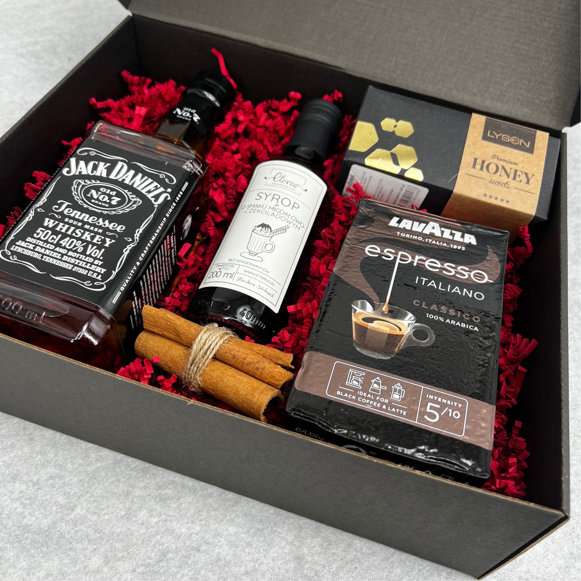 Elegancki, czarny box prezentowy wypełniony czerwonym konfetti, zawierający butelkę whisky Jack Daniels, syrop o smaku migdałowo-czekoladowym marki Eterno, opakowanie miodu Lyson, paczkę kawy...
