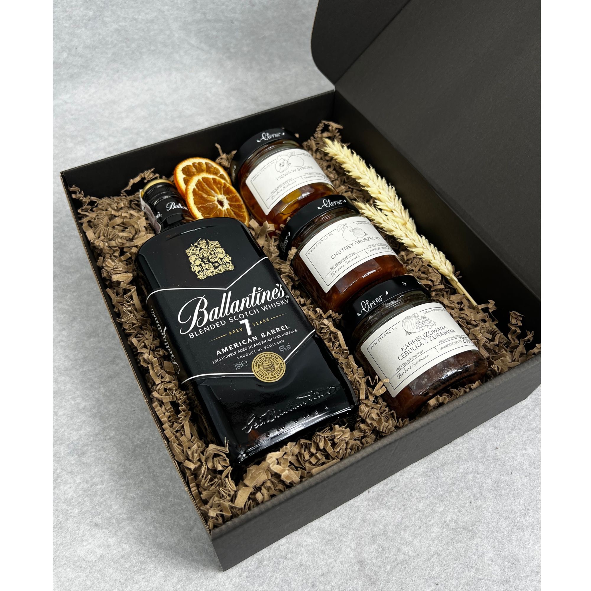 Czarny kosz prezentowy z whisky Ballantine's, suszonymi pomarańczami, trzema słoiczkami dżemów smakowych od Eterno i dekoracyjnym siankiem pszenicznym.
