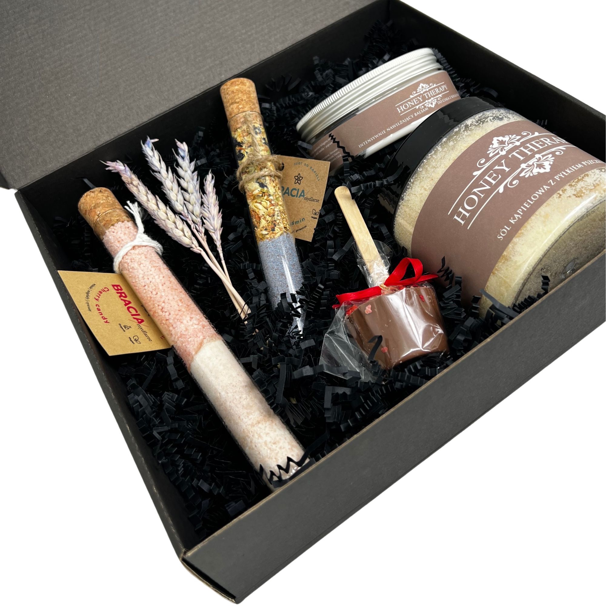 Czarny kartonowy box wypełniony czarnym papierem, zawierający zestaw produktów do pielęgnacji i relaksu: dwie szklane probówki z korkiem, wypełnione kolorowymi solami do kąpieli, mały słoiczek...