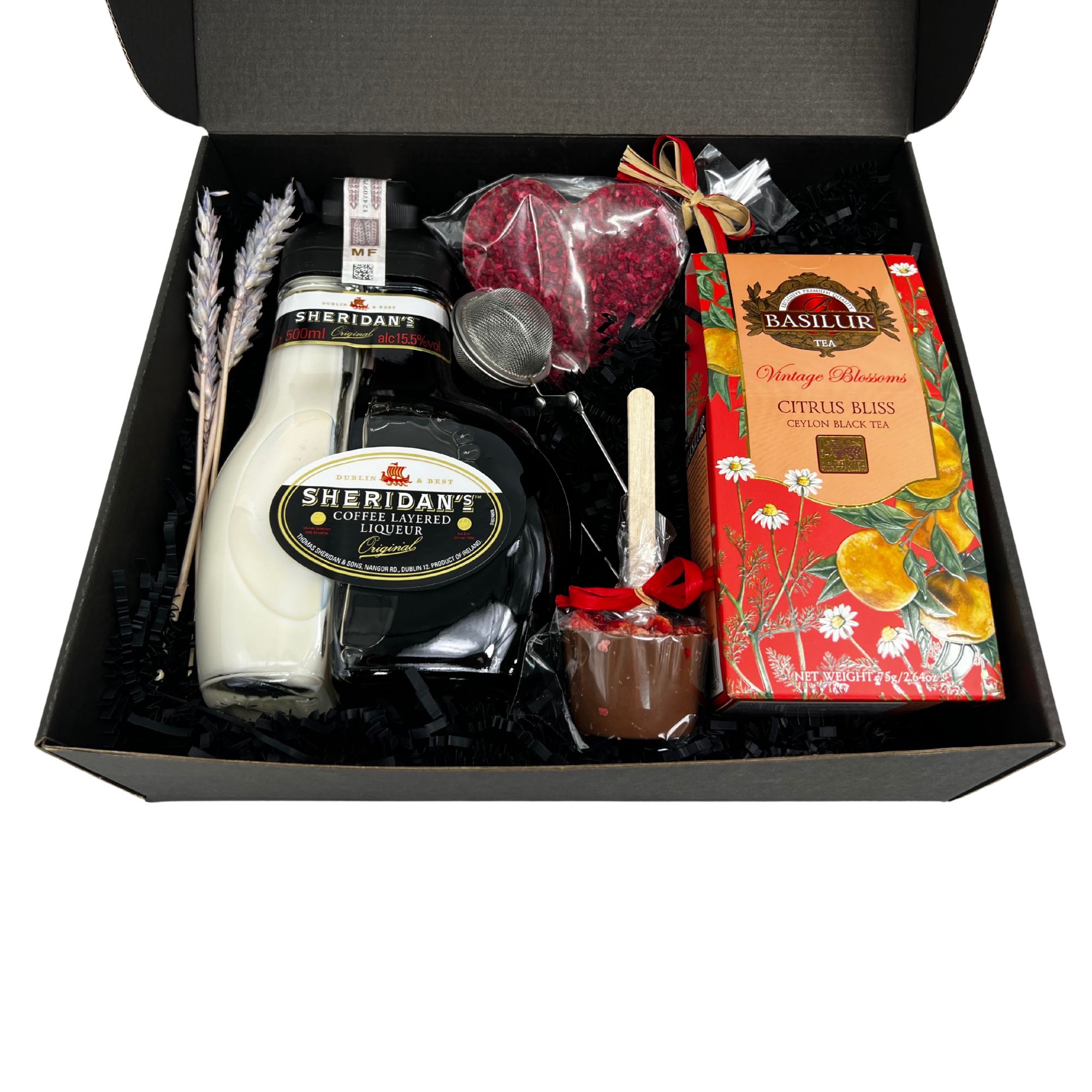 Czarny pudełko prezentowe wypełnione czarnym wypełniaczem, zawierające butelkę likieru kawowego Sheridan's, opakowanie herbaty Basilur Citrus Bliss, czekoladowy kubek z patyczkiem, serce z owoców...
