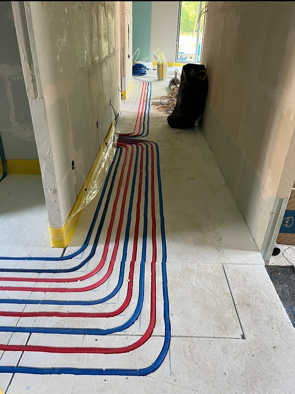 Układanie czerwonych i niebieskich rur grzewczych pod podłogą w trakcie prac instalacyjnych, widoczne także materiały budowlane i narzędzia.