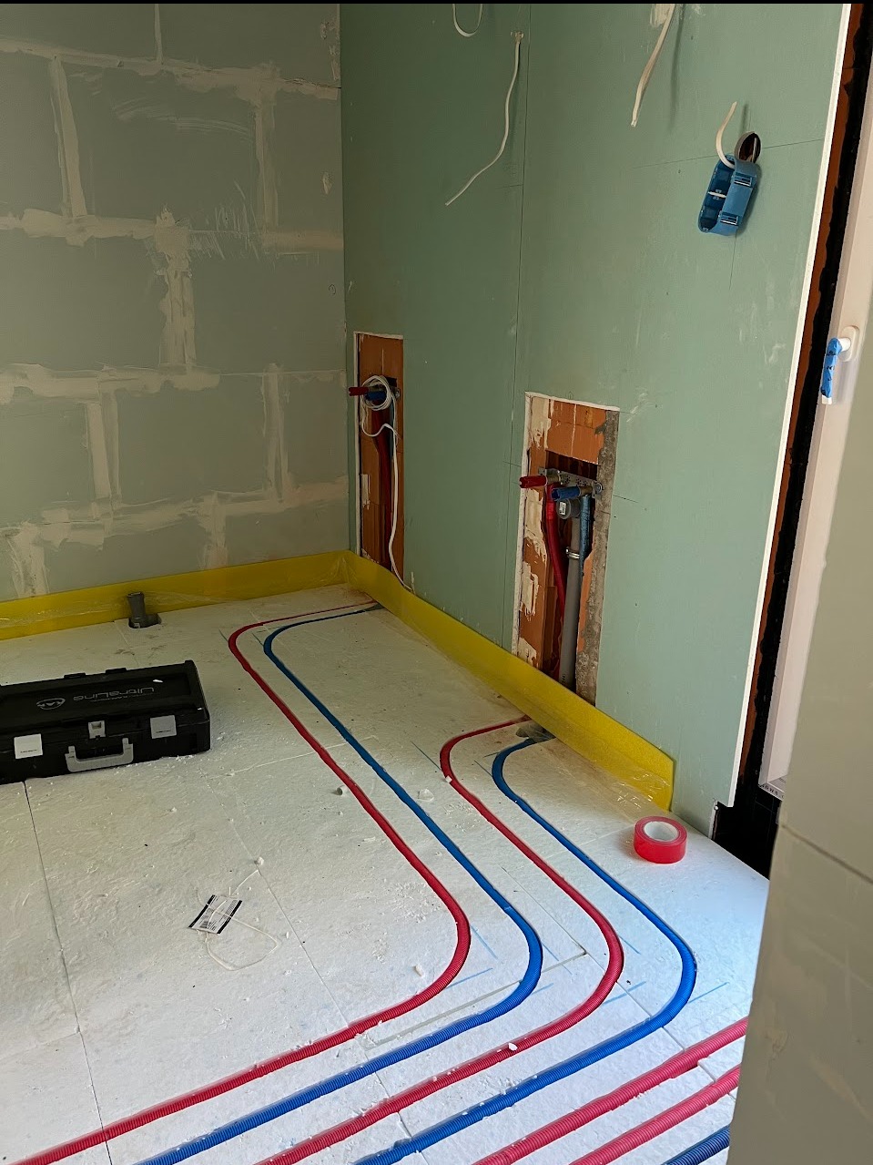 Wnętrze w trakcie instalacji ogrzewania podłogowego: widoczne rury grzewcze w kolorze czerwonym i niebieskim ułożone na izolacji, skrzynka narzędziowa obok ściany zabezpieczonej żółtą folią, ściany...