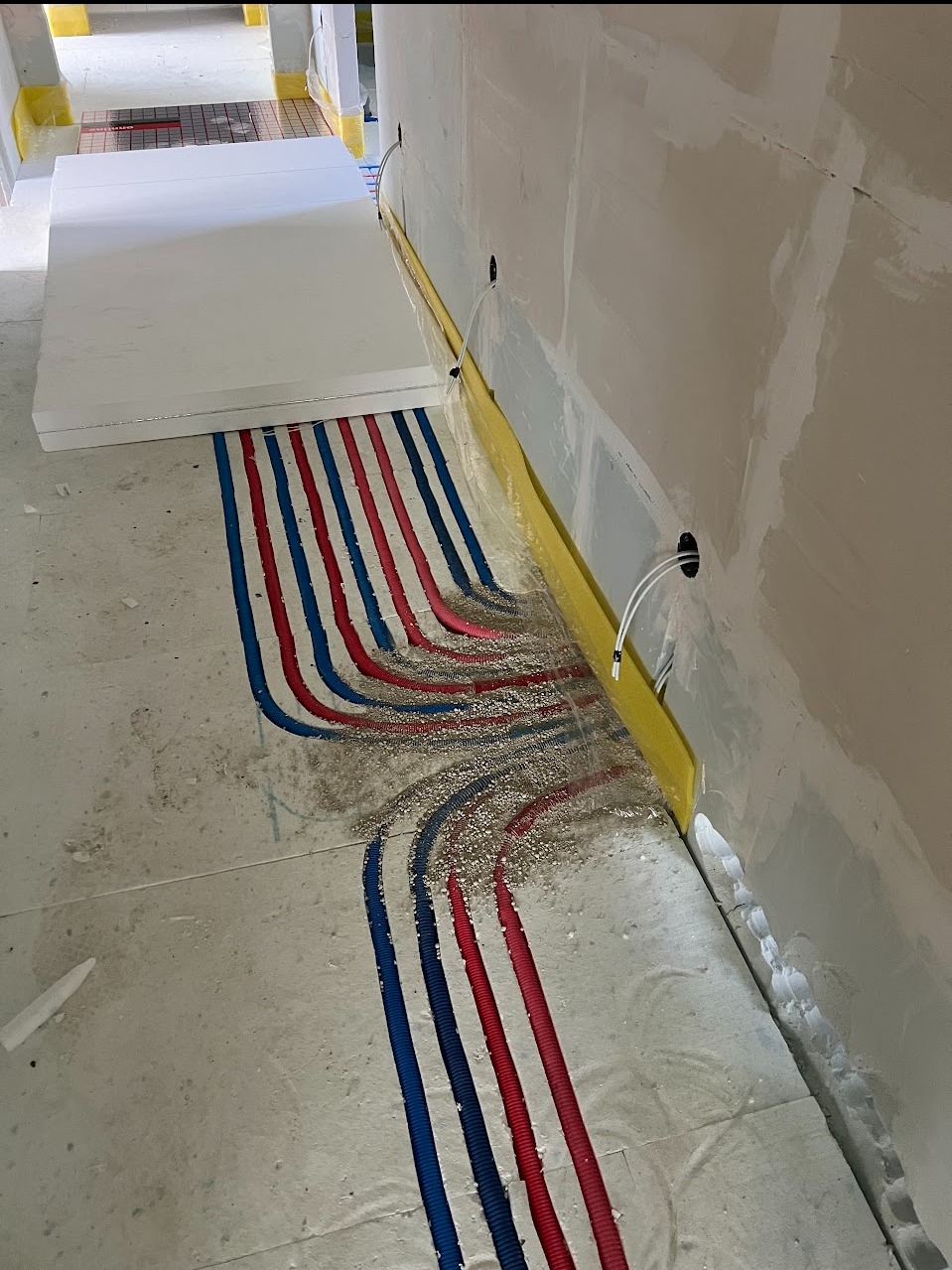 Instalacja ogrzewania podłogowego: czerwone i niebieskie rury prowadzone w wylewce, częściowo zasypane piaskiem, widoczne płyty izolacyjne i folia zabezpieczająca ściany.