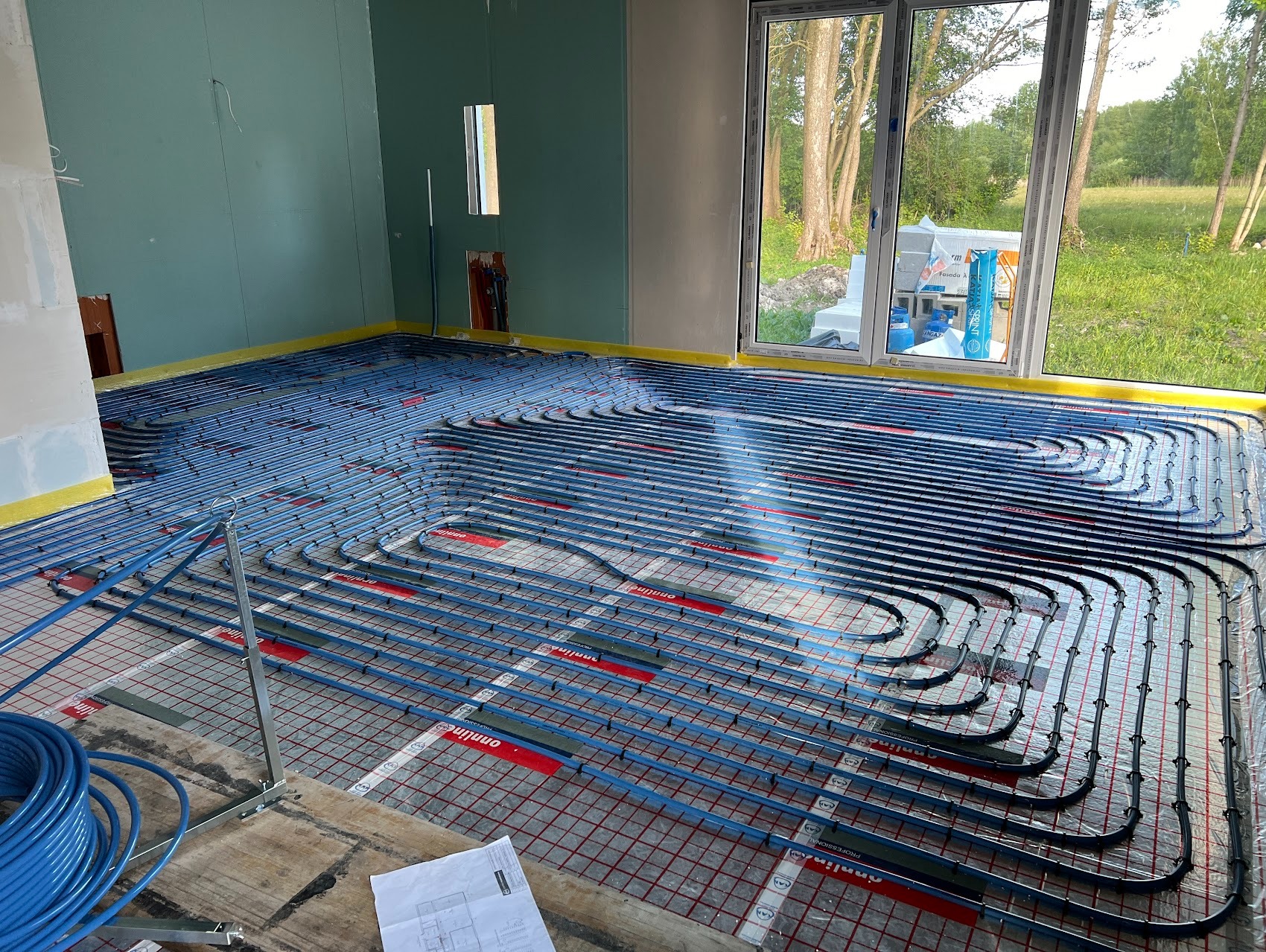 Instalacja ogrzewania podłogowego z niebieskich rur na siatce metalowej w surowym wnętrzu, widok z bliska na rury i rozłożony plan pomieszczenia.