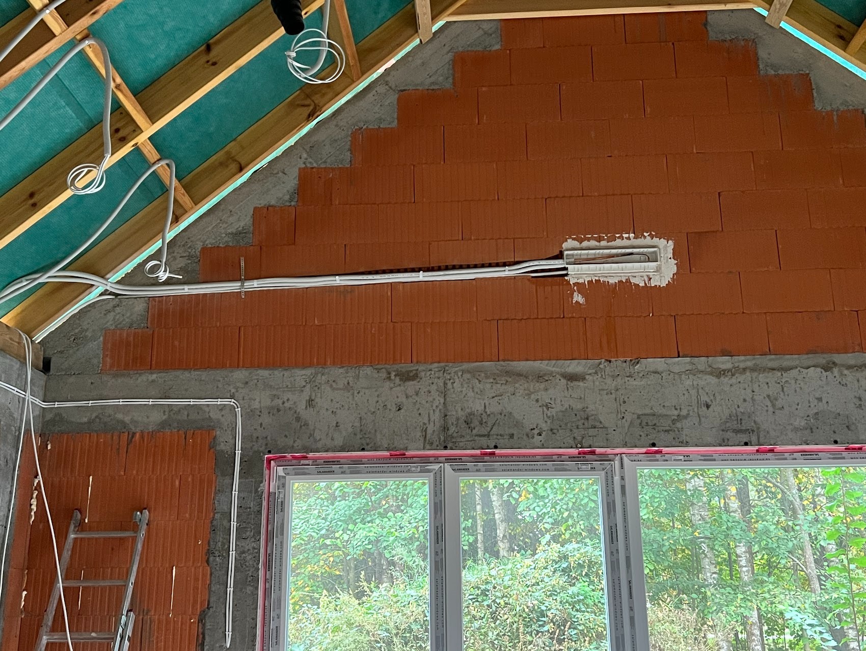 Wnętrze w trakcie budowy: ściana z czerwonej cegły, widoczne przewody elektryczne prowadzone po ścianie, okno PCV z widokiem na las, drabina oparta o ścianę.