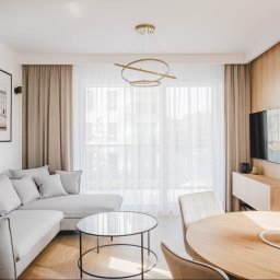 Moya Stolarnia Krzysztof Zawadzki - Jasne wnętrze salonu z szarą sofą, okrągłym stolikiem kawowym, drewnianym stołem jadalnym i telewizorem na ścianie obłożonej drewnianą okładziną. Na ścianie wiszą grafiki w czarnych ramach.