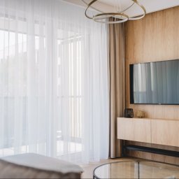 Moya Stolarnia Krzysztof Zawadzki - Nowoczesny salon z drewnianą ścianą, telewizorem, wiszącą szafką RTV, delikatnymi firanami i okrągłym szklanym stolikiem kawowym.