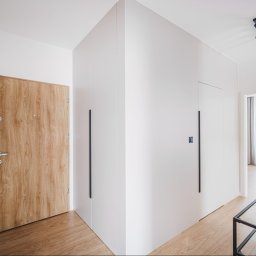Moya Stolarnia Krzysztof Zawadzki - Nowoczesny przedpokój z białą zabudową szaf wnękowych z długimi, pionowymi uchwytami i drewnianymi drzwiami wejściowymi, oświetlony trzypunktowym reflektorem sufitowym.