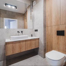 Moya Stolarnia Krzysztof Zawadzki - Nowoczesna łazienka z drewnianą zabudową szafek pod umywalką, wiszącą toaletą i grzejnikiem drabinkowym, oświetlona punktowymi lampami i wiszącymi lampami tubowymi.