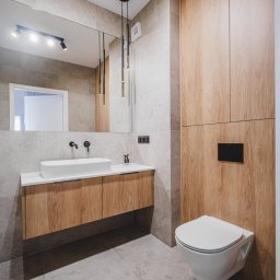 Moya Stolarnia Krzysztof Zawadzki - Nowoczesna łazienka z drewnianą szafką pod umywalką, wiszącą toaletą i szarą płytką na ścianach i podłodze, oświetlona wiszącą lampą z elementami złota.