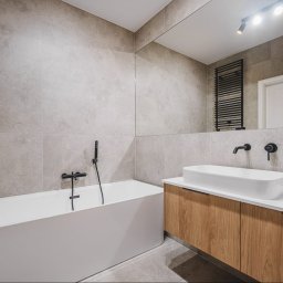 Moya Stolarnia Krzysztof Zawadzki - Nowoczesna łazienka z wanną, umywalką nablatową na drewnianej szafce, szarą płytką ścienną i czarną armaturą, oświetlona punktowymi lampami sufitowymi.