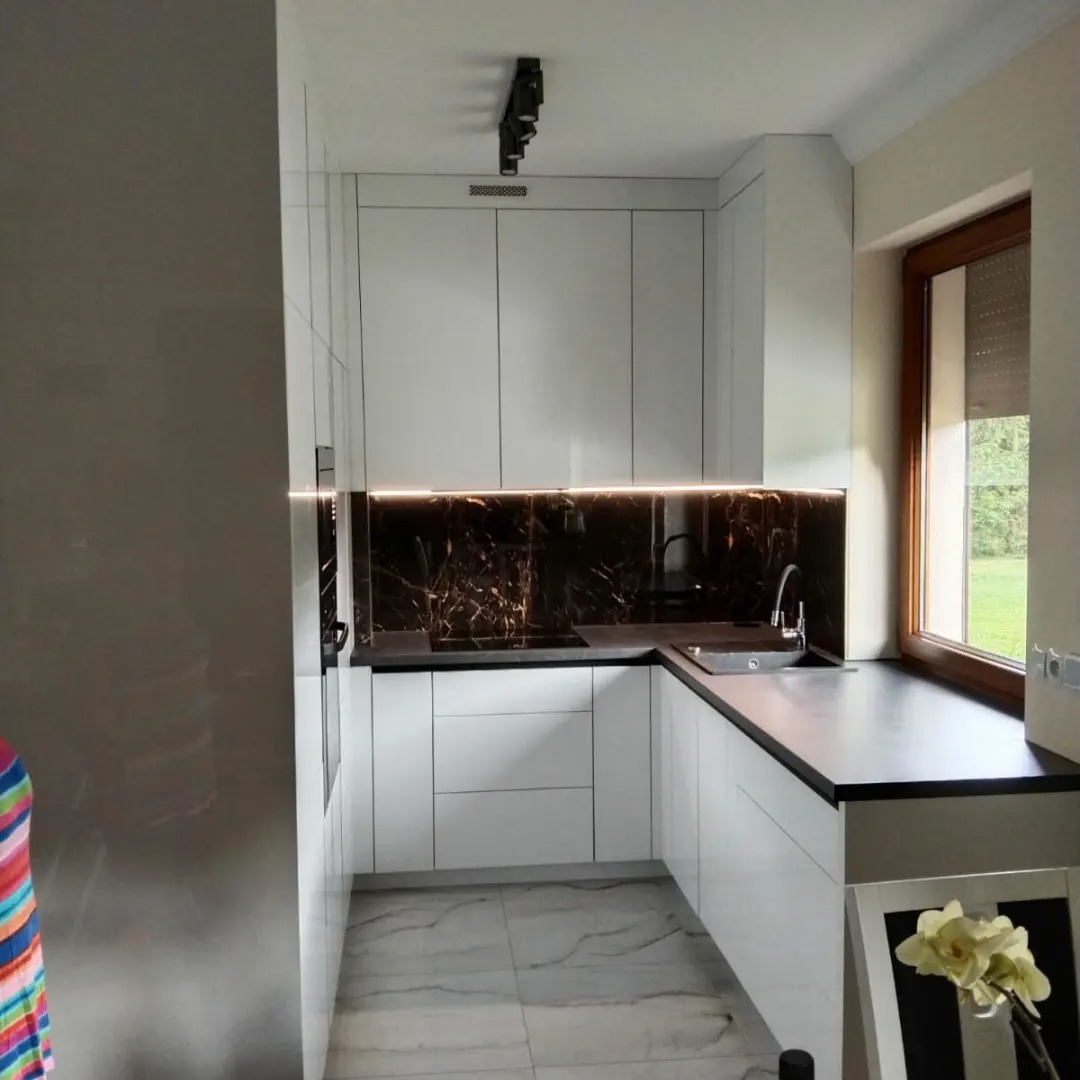 Nowoczesna kuchnia z białymi frontami, czarnym blatem i marmurowym panelem nad blatem, oświetlona listwą LED, z widokiem na zieleń za oknem.