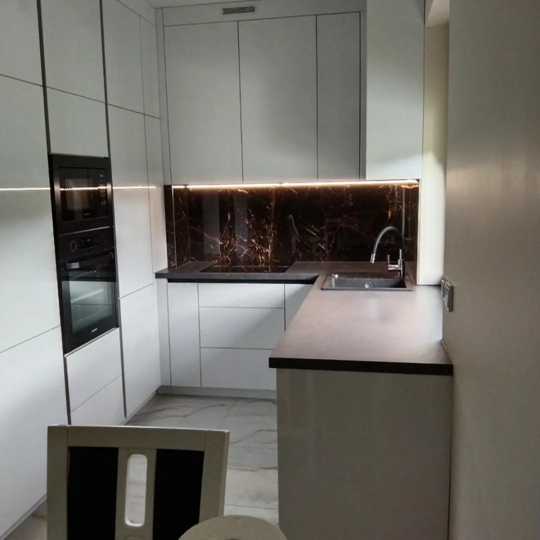 Nowoczesna kuchnia z białymi, lakierowanymi frontami, czarnym blatem i marmurowym panelem nad blatem, oświetlonym listwą LED.