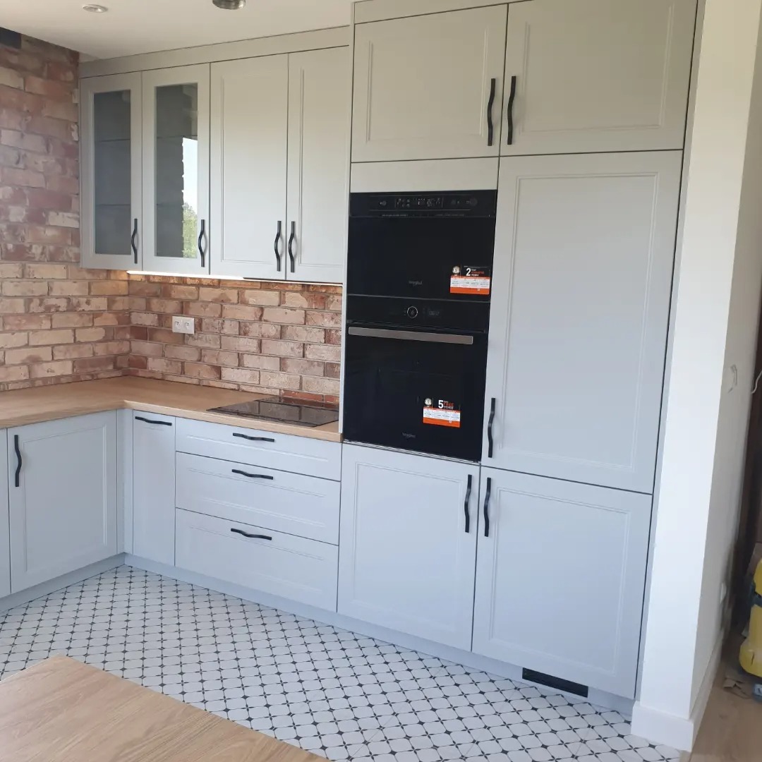 Jasnoszara zabudowa kuchenna z piekarnikiem, drewnianym blatem i ceglaną ścianą w tle, uzupełniona wzorzystą podłogą.