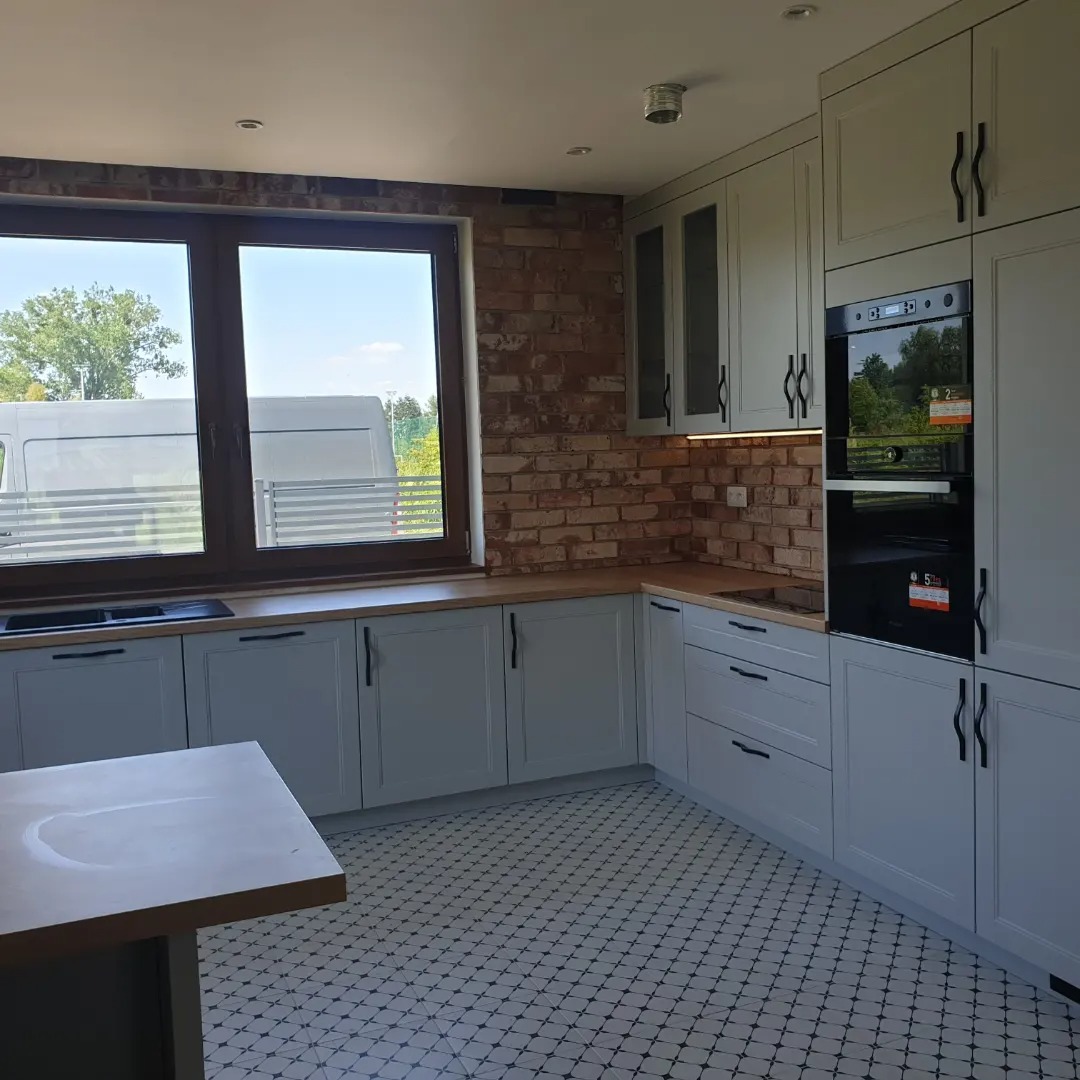 Jasnoszara zabudowa kuchenna z ceglanym backsplashiem w Ciechocinie, widok na okno z białym busem na zewnątrz, wzorzysta podłoga.