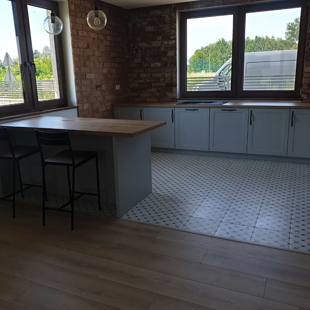 Kuchnia w stylu loft z ceglaną ścianą, jasnoszarymi szafkami, drewnianym blatem i wyspą z dwoma czarnymi hokerami, podłoga wyłożona wzorzystymi płytkami.