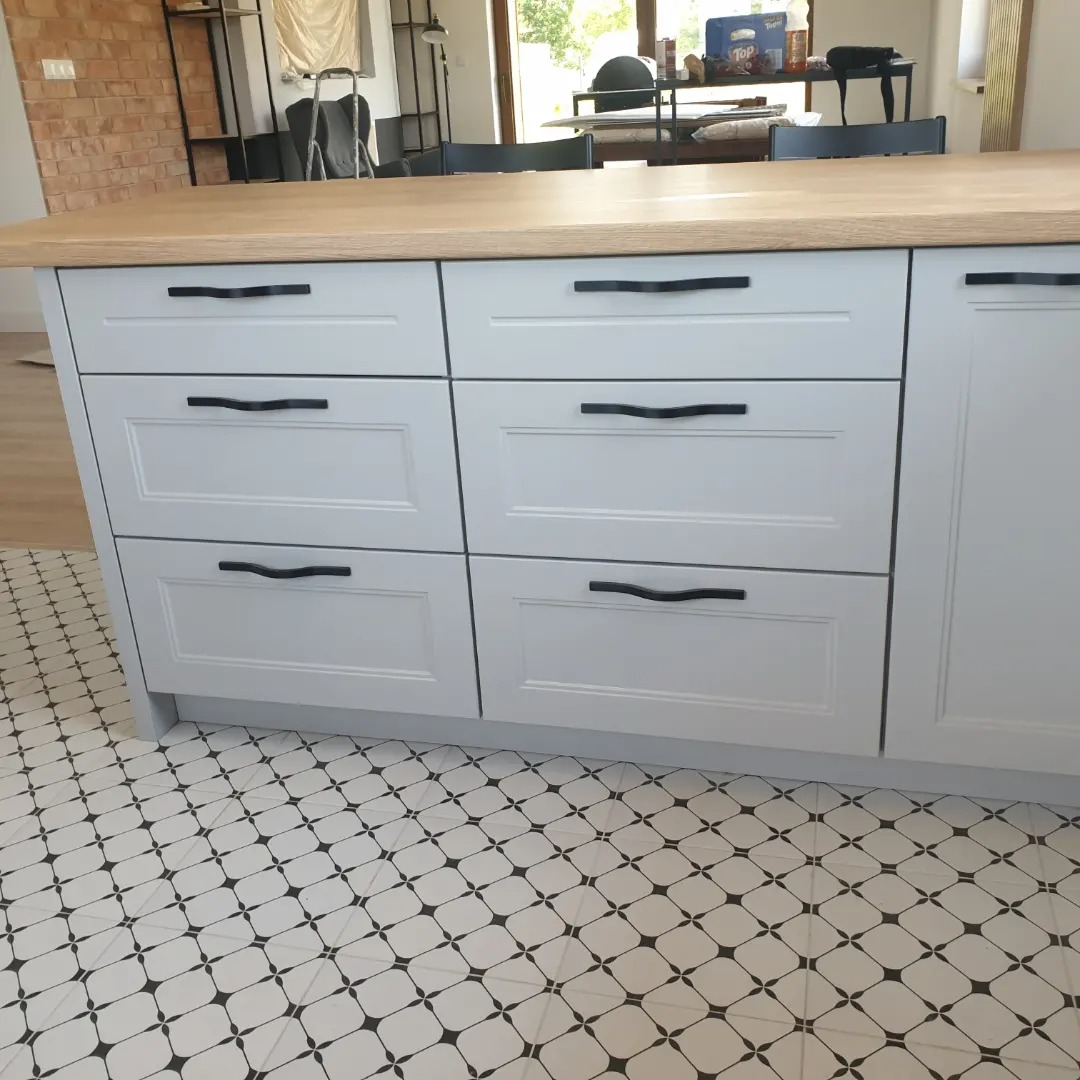 Jasnoszare szafki kuchenne z drewnianym blatem i czarnymi uchwytami na tle wzorzystej podłogi.