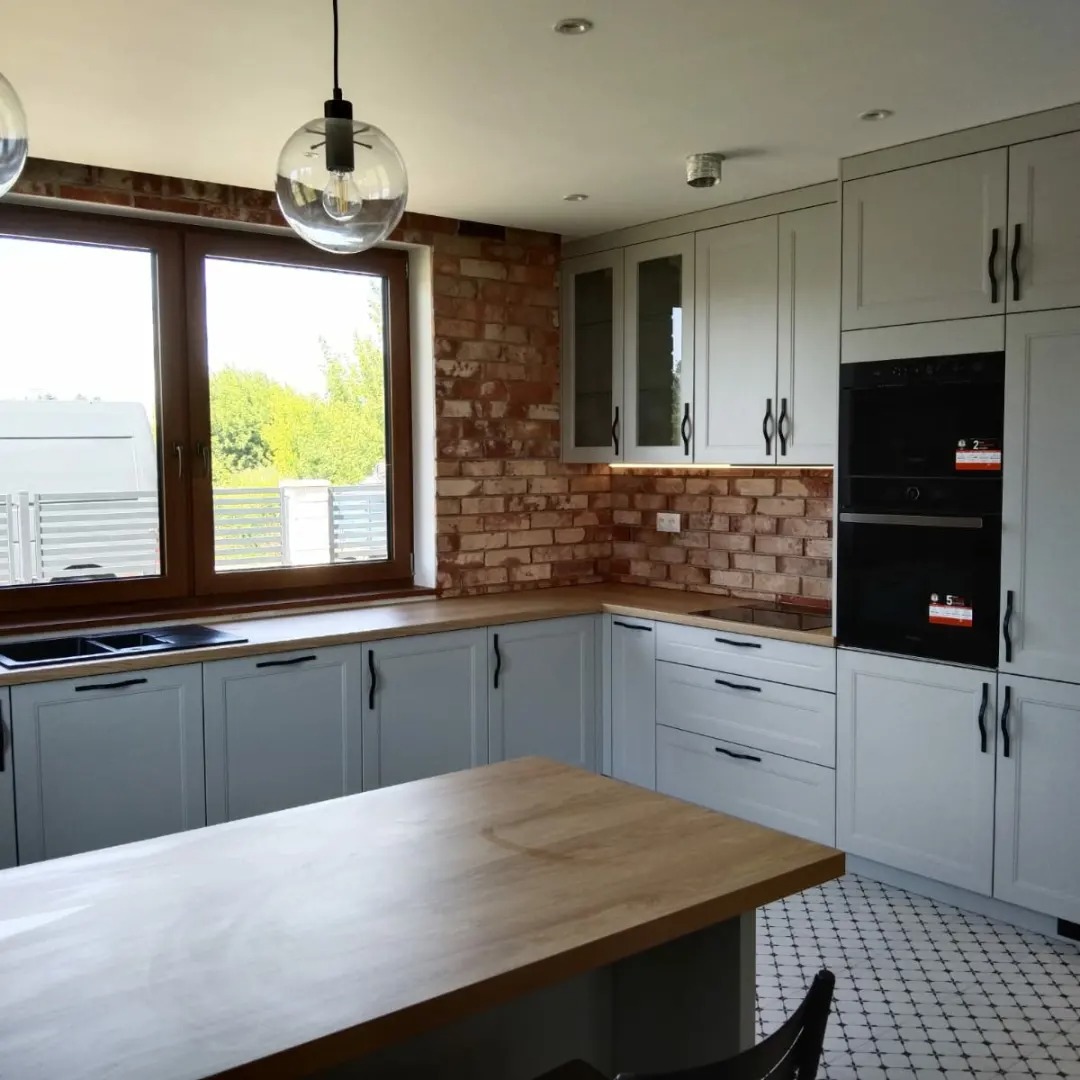 Jasna kuchnia z szarymi szafkami, drewnianym blatem i ceglaną ścianą w tle, oświetlona wiszącą lampą z przezroczystym kloszem.