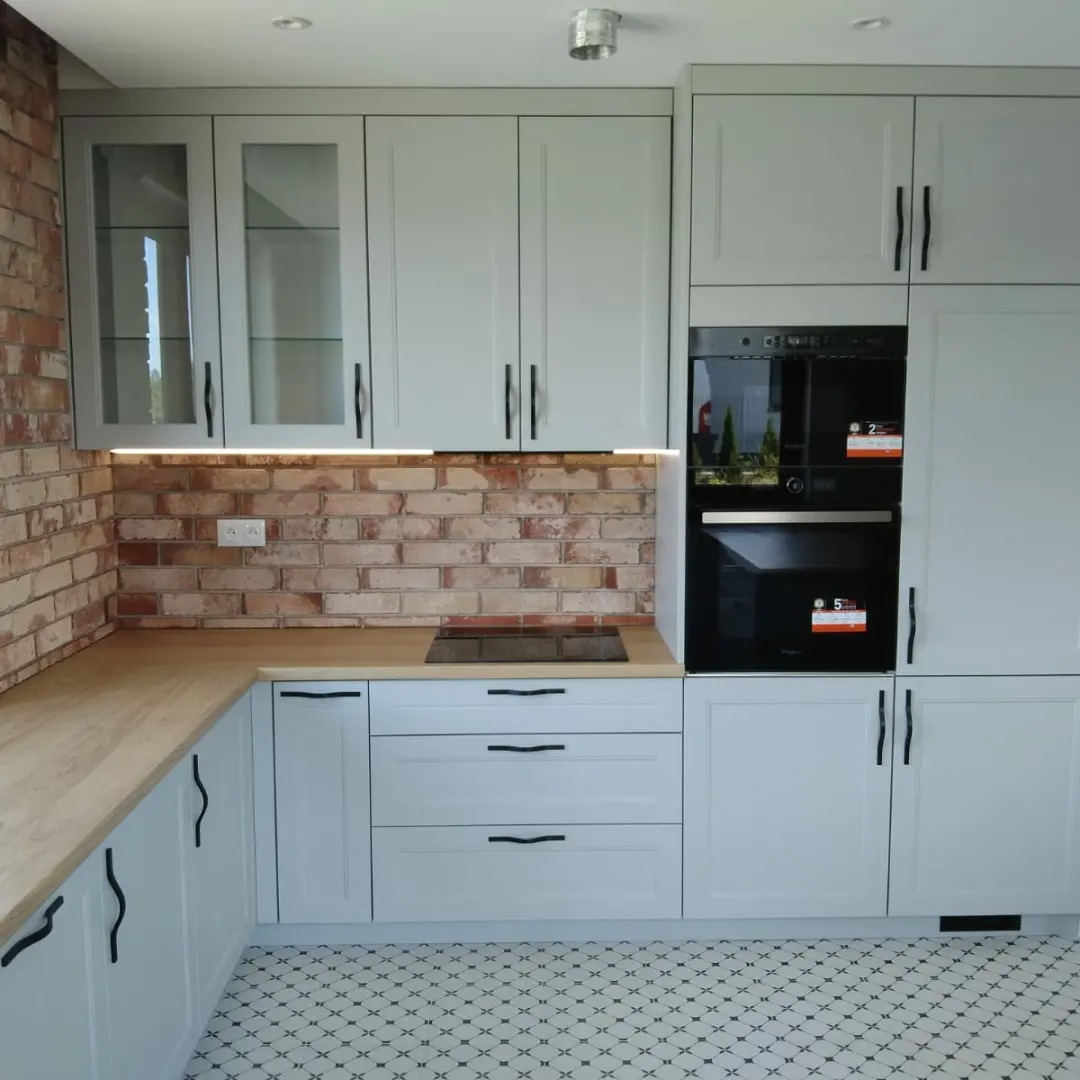 Jasnoszara kuchnia na wymiar z ceglaną ścianą, drewnianym blatem i czarnymi uchwytami, uzupełniona o czarny piekarnik w zabudowie i wzorzystą podłogę.