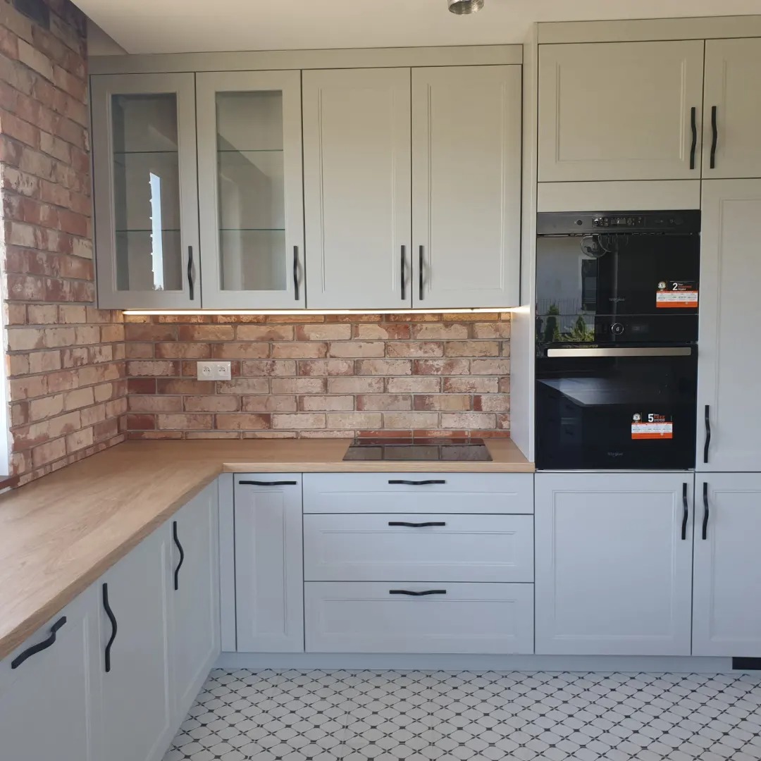 Jasnoszara zabudowa kuchenna z drewnianym blatem i ceglaną ścianą, oświetlenie LED pod szafkami, czarny piekarnik w zabudowie i wzorzysta podłoga.