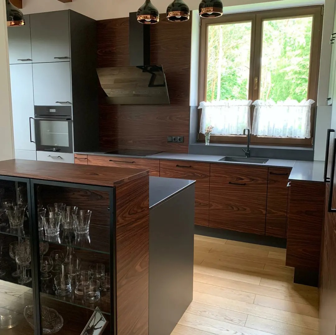 Nowoczesna kuchnia z drewnianymi frontami w odcieniach brązu, szare szafki górne, czarny okap i wyspa, szklana witryna z kryształami i parkiet w jodełkę.