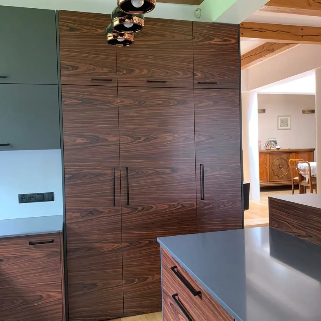 Nowoczesna kuchnia z zabudową sięgającą sufitu, fronty z ciemnego drewna o wyraźnym usłojeniu, czarne, minimalistyczne uchwyty, szare blaty i szafki po lewej stronie, widok na jadalnię z drewnianym...