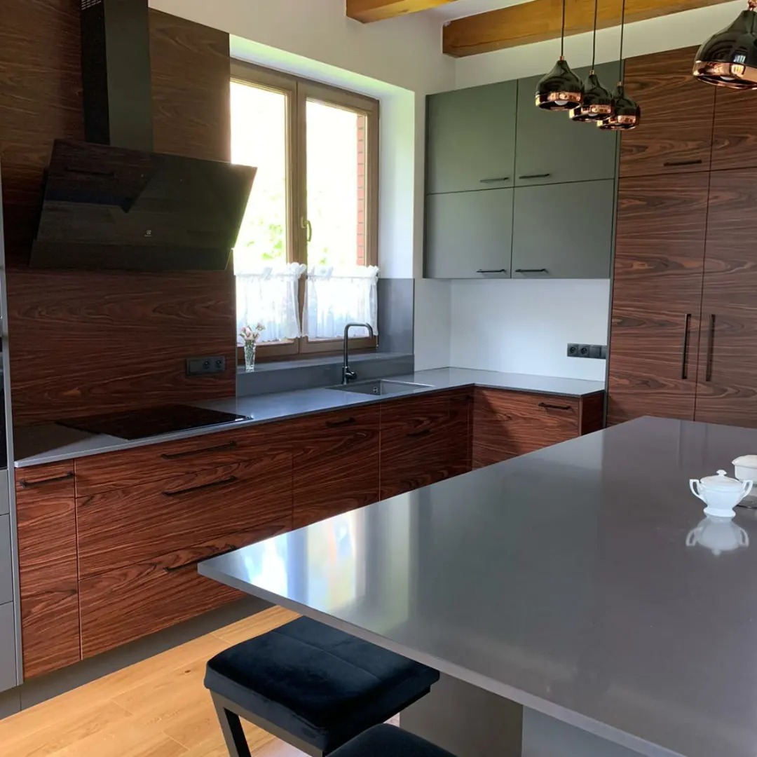 Nowoczesna kuchnia z ciemnego drewna i szarych szafek, z wyspą kuchenną i czarnymi lampami wiszącymi, widok na okno z firankami.