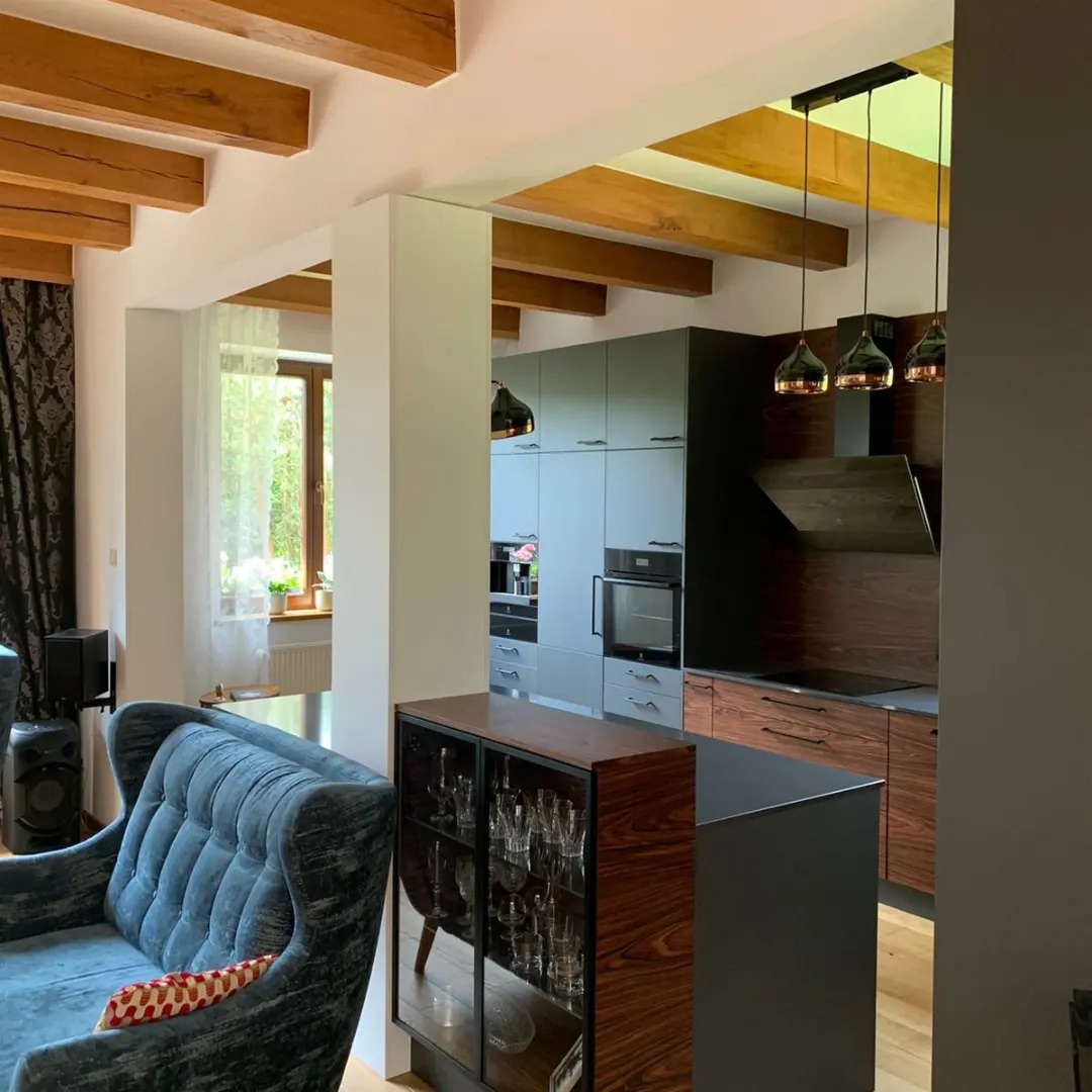 Nowoczesna kuchnia z szarymi szafkami, drewnianym blatem i wyspą, widoczna z perspektywy salonu z błękitnym fotelem i drewnianymi belkami stropowymi.