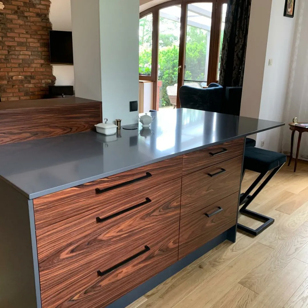 Wyspa kuchenna z ciemnobrązowymi frontami szuflad o wyraźnym usłojeniu, szarym blatem i czarnymi uchwytami, uzupełniona o białą porcelanę i metalowe akcesoria na blacie, z widokiem na salon...