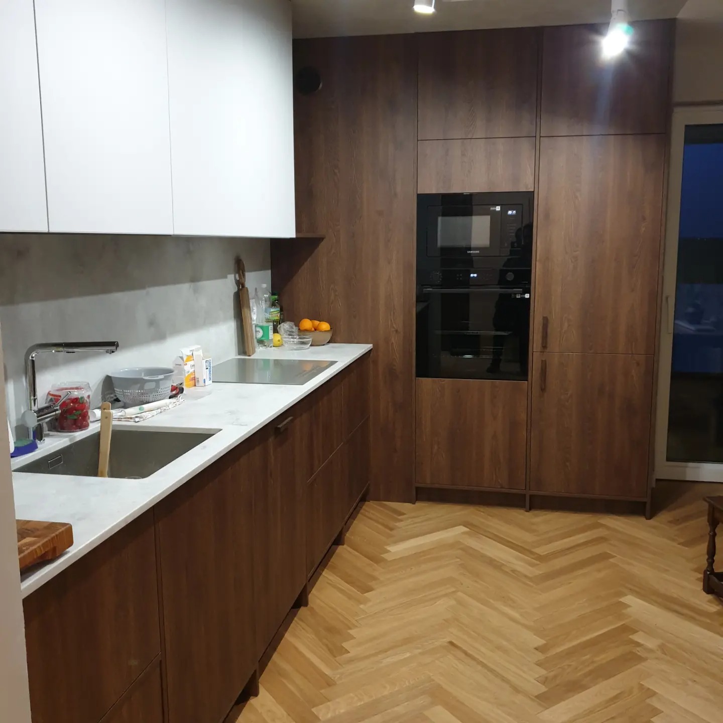 Nowoczesna kuchnia z drewnianymi frontami szafek dolnych i białymi górnymi, marmurowy blat, parkiet ułożony w jodełkę, zabudowany sprzęt AGD.