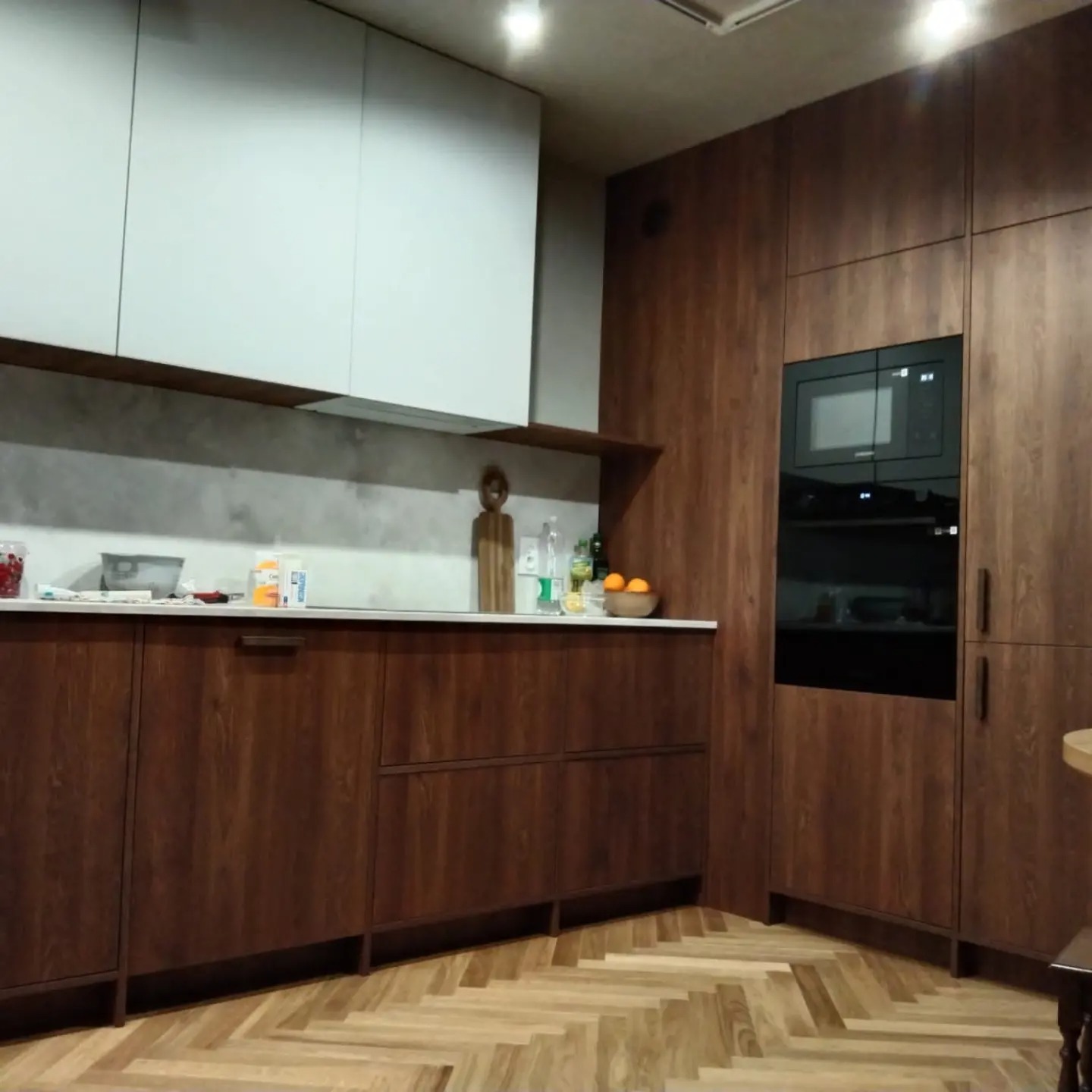 Nowoczesna kuchnia z drewnianymi frontami szafek, białymi szafkami górnymi i parkietem ułożonym w jodełkę, z wbudowanym piekarnikiem i mikrofalówką.