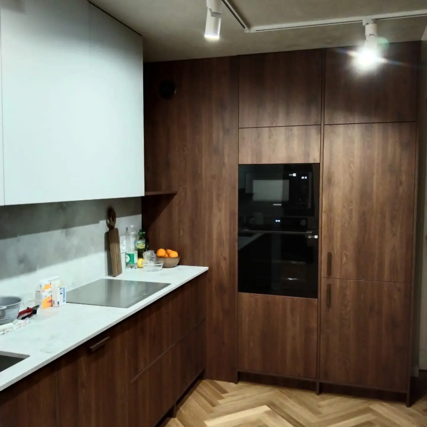 Nowoczesna kuchnia z drewnianymi frontami, białymi górnymi szafkami i marmurowym blatem, oświetlona punktowymi lampami sufitowymi, z parkietem ułożonym w jodełkę.