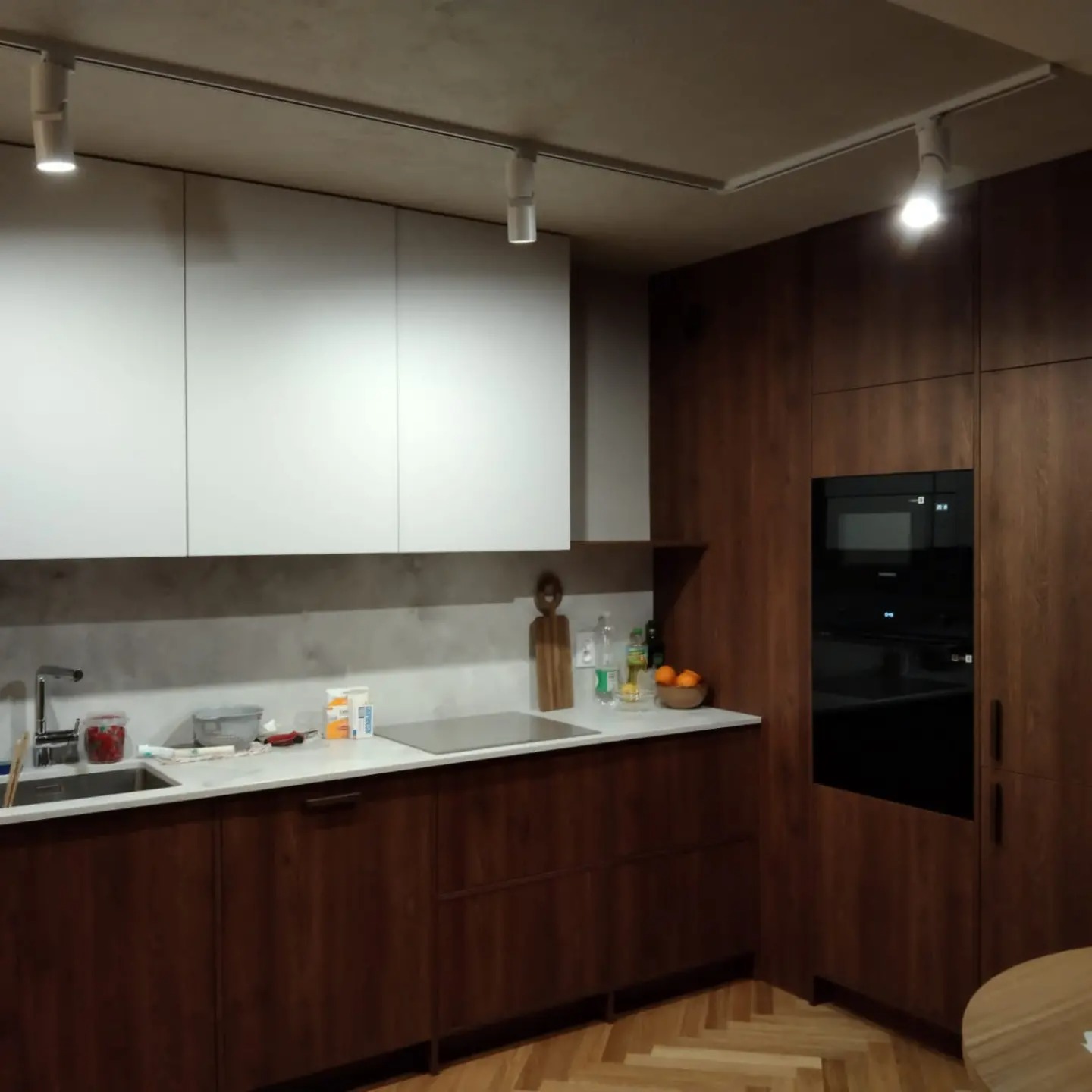 Nowoczesna kuchnia z białymi szafkami górnymi i drewnianymi szafkami dolnymi, oświetlenie punktowe na szynoprzewodzie, zabudowany piekarnik, jasny blat i parkiet w jodełkę.