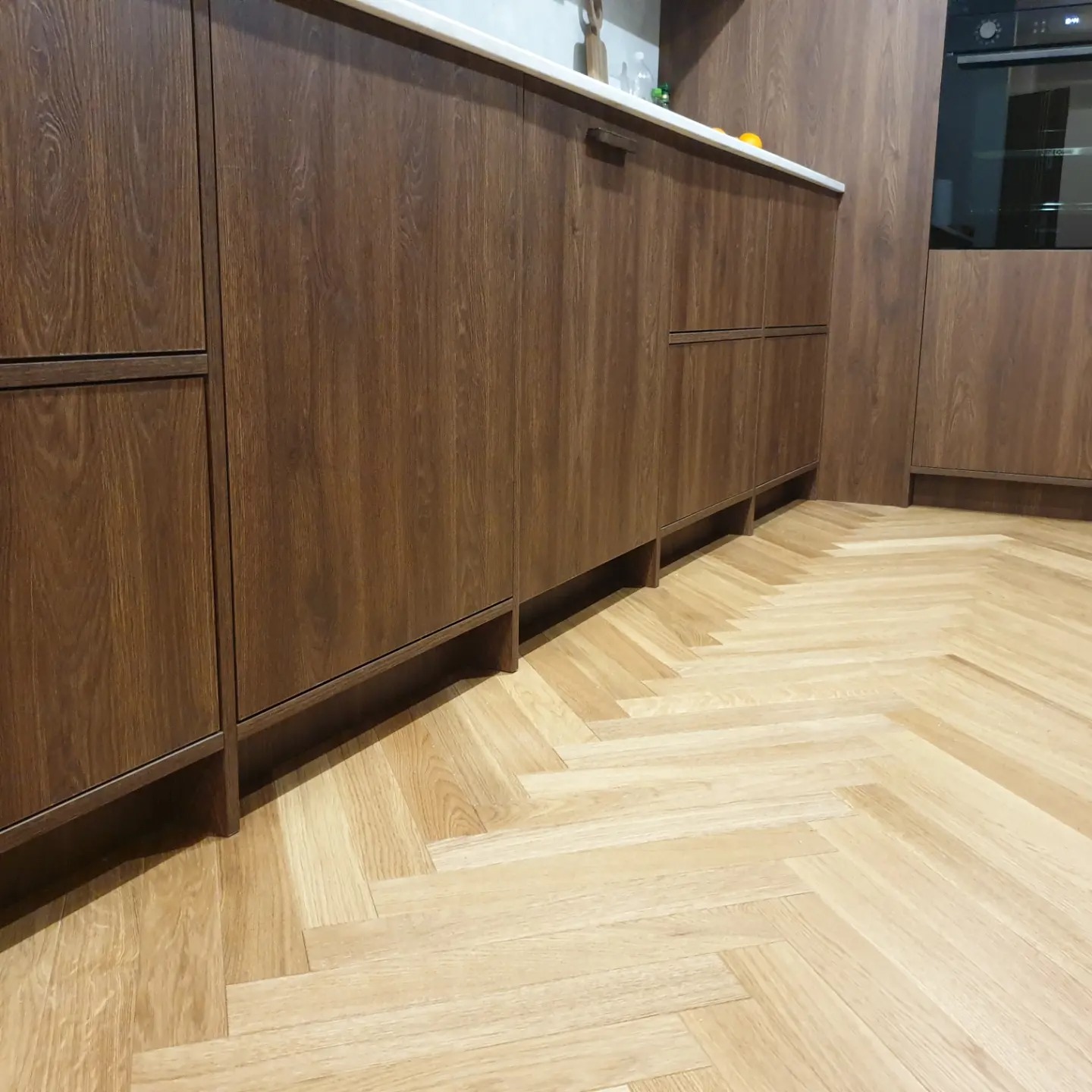 Ciemnobrązowe, drewnopodobne szafki kuchenne z białym blatem i parkietem w jodełkę na podłodze, widok z niskiej perspektywy.