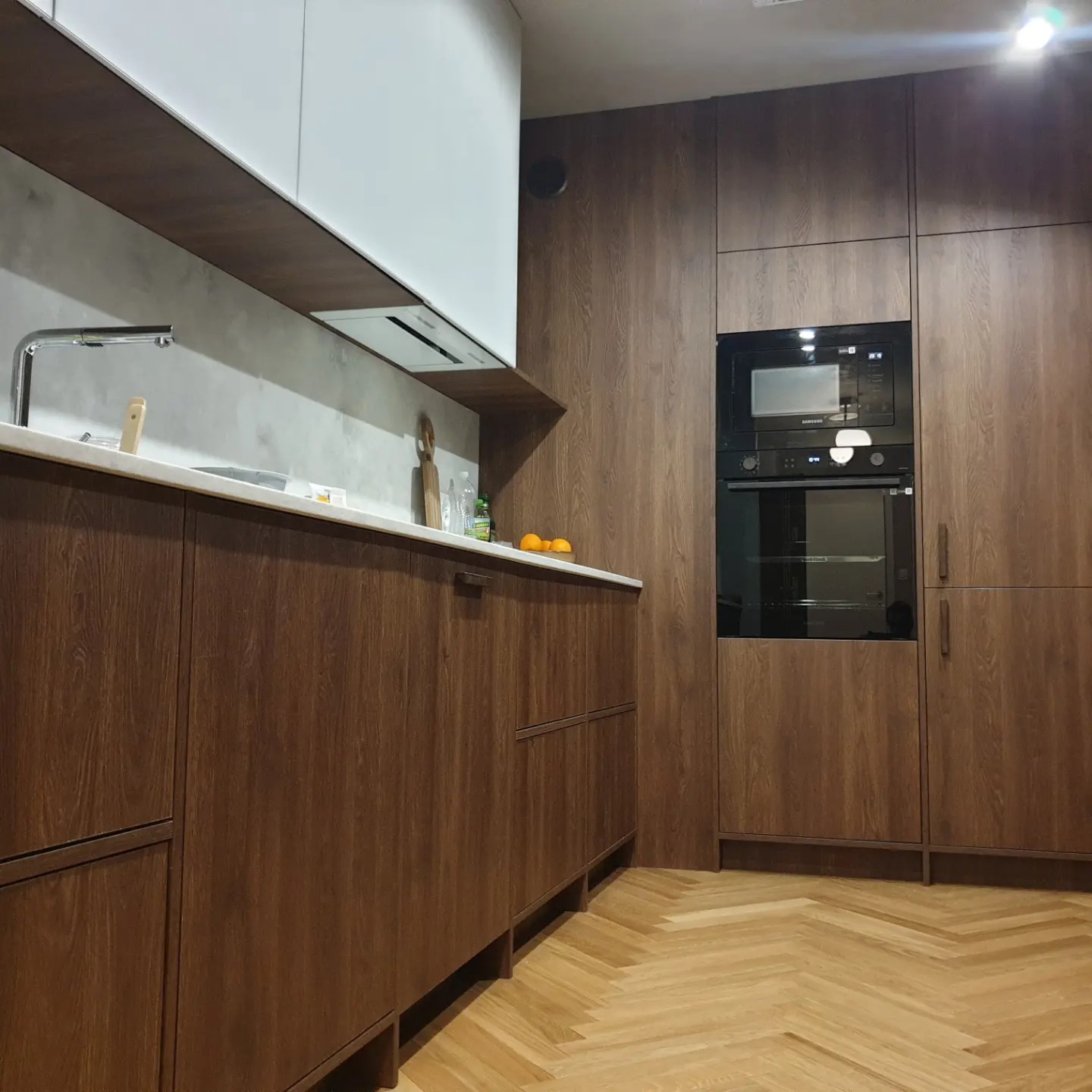 Nowoczesna kuchnia z drewnianymi frontami szafek, białymi szafkami górnymi i parkietem w jodełkę. W zabudowie piekarnik i mikrofalówka.