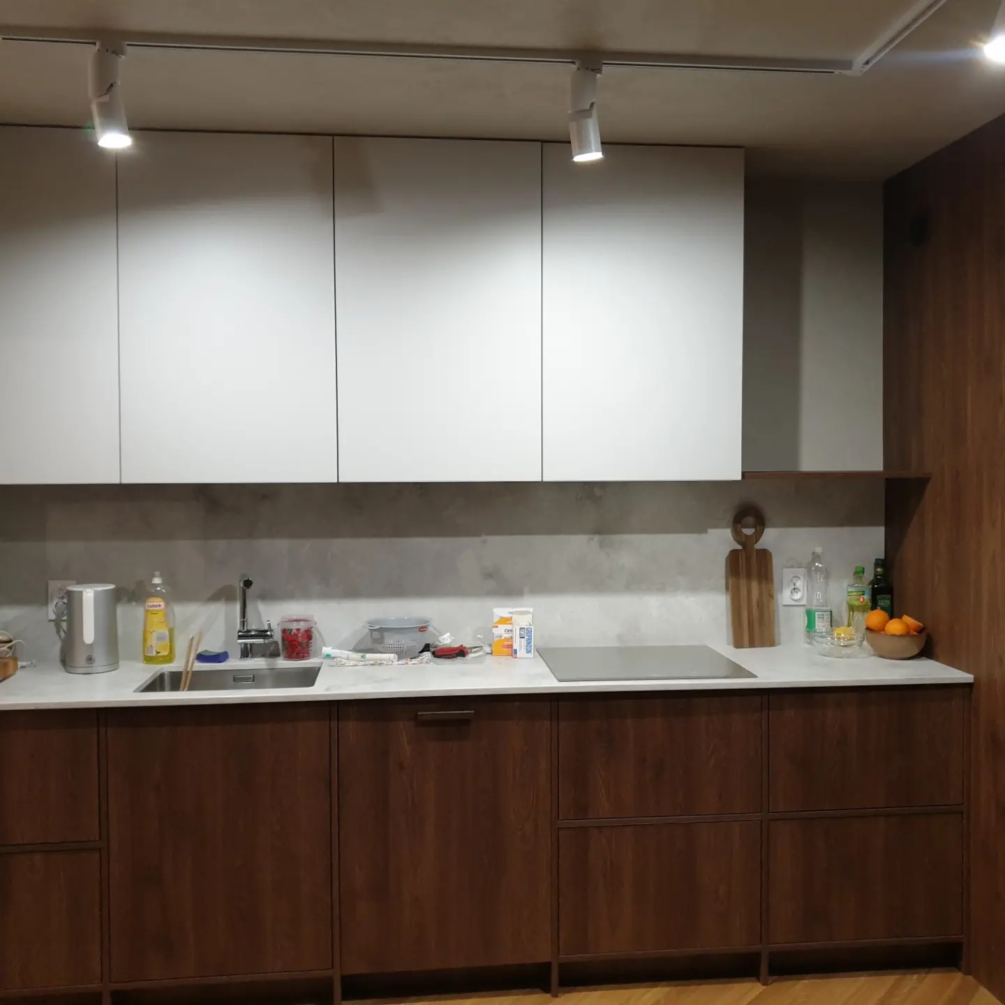 Nowoczesna kuchnia: dolne szafki z ciemnego drewna, białe górne szafki podświetlone reflektorami, blat z jasnego kamienia, widoczne akcesoria kuchenne i owoce.