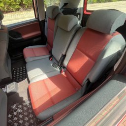 Łukasz Rusiński LukClean - Wnętrze samochodu z trzema rzędami siedzeń w odcieniach szarości i czerwieni, widoczne pasy bezpieczeństwa i gumowa wycieraczka pod nogami pasażerów.
