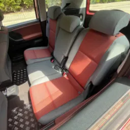 Wnętrze samochodu z trzema rzędami siedzeń w odcieniach szarości i czerwieni, widoczne pasy bezpieczeństwa i gumowa wycieraczka pod nogami pasażerów.