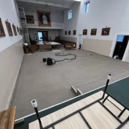 Duża sala z dywanem w trakcie czyszczenia, widoczny odkurzacz piorący i wiązka kabli na dywanie, w tle ściany z obrazami i schody, w rogu sali ustawione stoły i ławki.