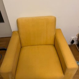 Łukasz Rusiński LukClean - Żółty fotel z plamami na siedzisku i oparciu, stojący na drewnianej podłodze przed białą ścianą, widoczne ślady po czyszczeniu.