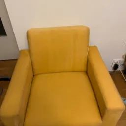 Żółty fotel z plamami na siedzisku i oparciu, stojący na drewnianej podłodze przed białą ścianą, widoczne ślady po czyszczeniu.