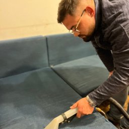 Łukasz Rusiński LukClean - Mężczyzna w okularach czyści niebieską kanapę za pomocą profesjonalnego odkurzacza z przezroczystą końcówką.
